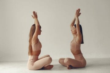 Two women in pink yoga outfits practicing yoga poses on mats in a bright, serene studio environment.
