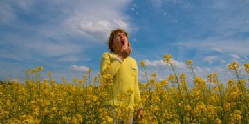 A man dressed in a yellow shirt stands surrounded by a sea of yellow flowers, experiencing allergy effects.