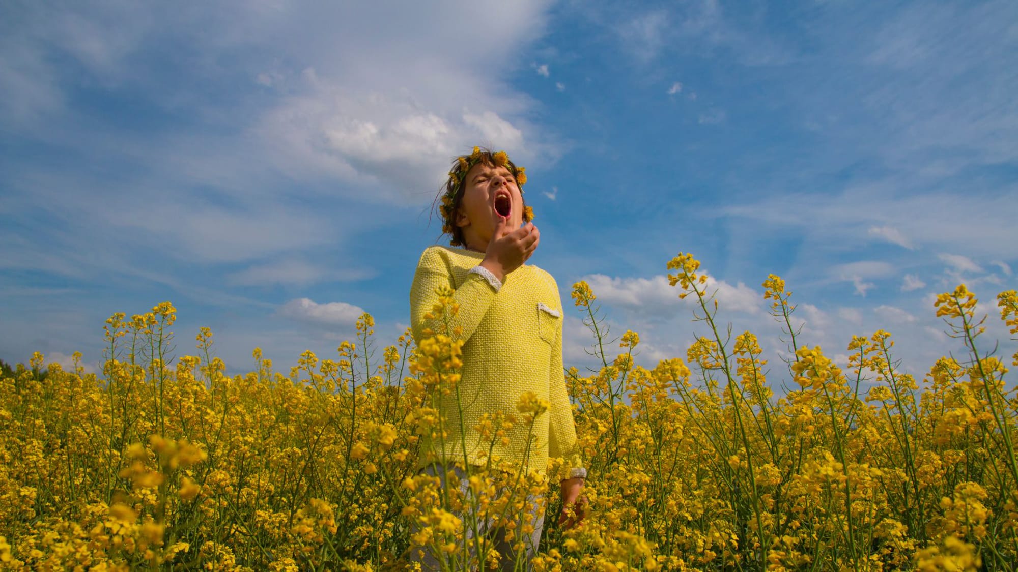 A man dressed in a yellow shirt stands surrounded by a sea of yellow flowers, experiencing allergy effects.
