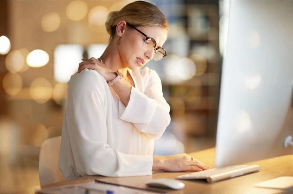 A woman winces in pain and puts her hand to her neck at her desk.